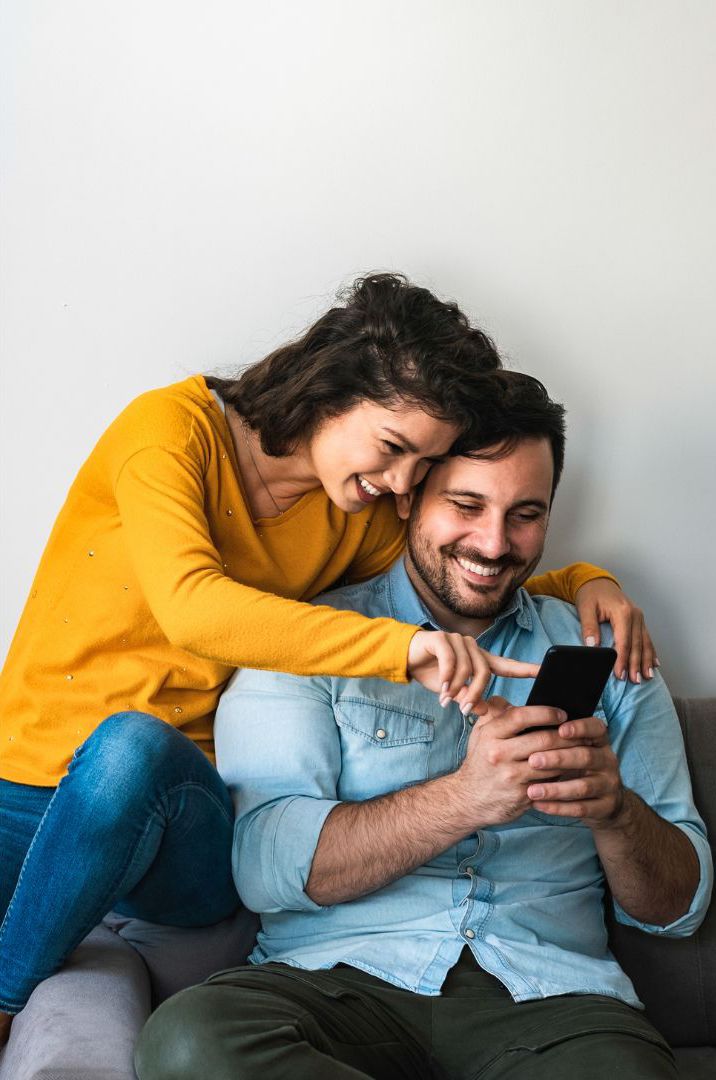 A woman wearing a thin mustard yellow sweatshirt and skinny jeans with curly hair is chatting with a man wearing a men's denim shirt and army green pants for men playing cell phones and laughing together on an aesthetic gray sofa in the living room