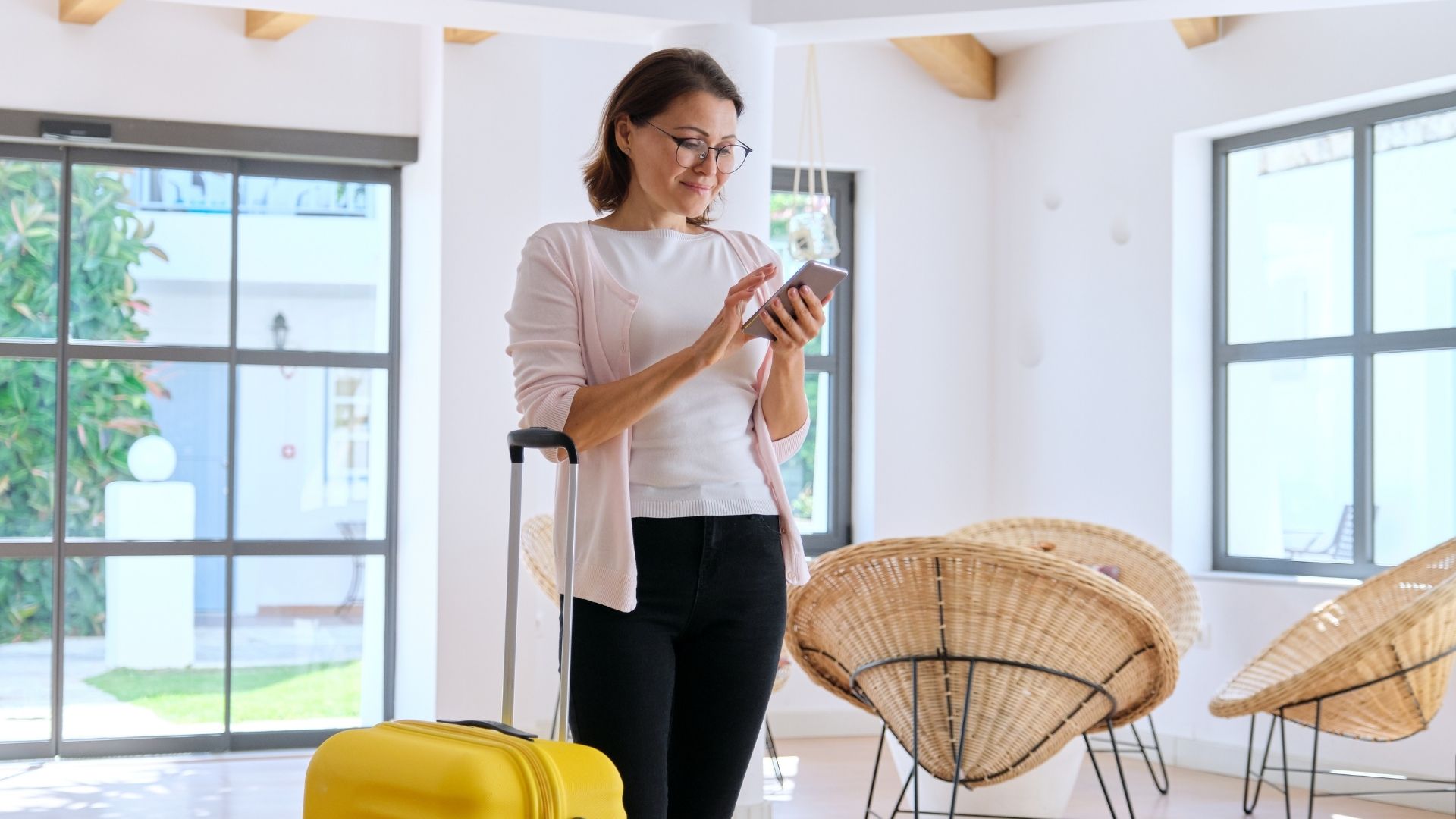 A woman with short brown hair wearing glasses is wearing a plain white t-shirt and light pink cardigan while carrying a yellow suitcase in a luxury hotel in Bali while looking for something on her cell phone about how to understand the target audience