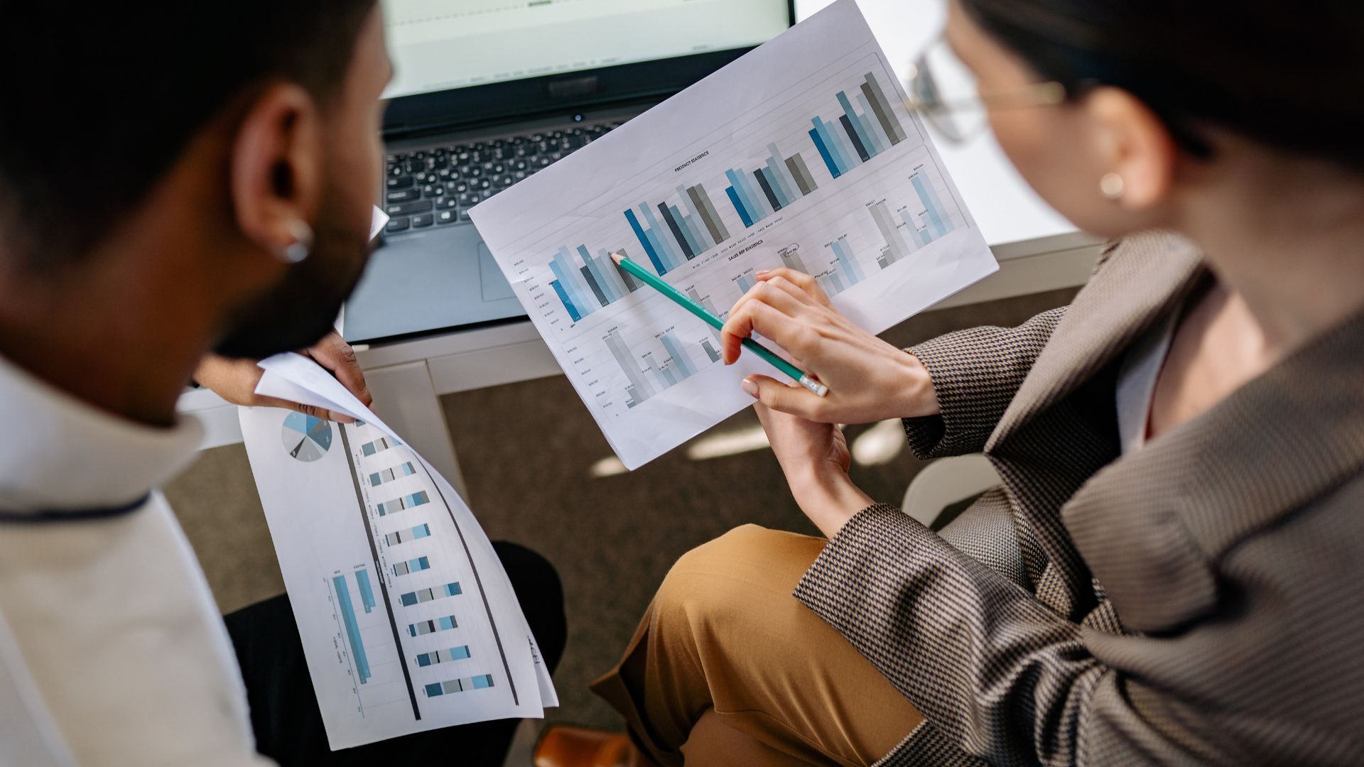 A woman wearing a white tanktop shirt covered with a brown patterned coat and brown casual formal office trousers wearing glasses is explaining about data and analytics to her colleague using HVS paper containing blue bar chart data whose colleague is a man wearing a white shirt and has a beard