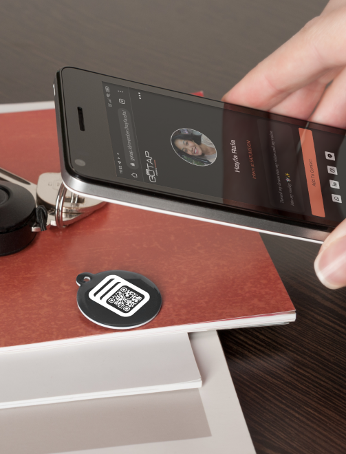 A smartphone showcasing the user interface of GOTAP, alongside a GOTAP NFC smart tag placed on a working table