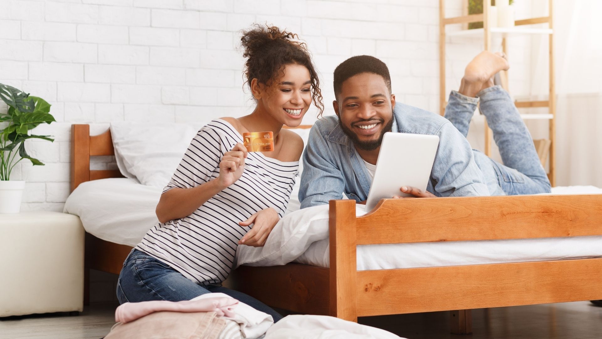 A woman wearing a black and white striped sabrina and skinny jeans with curly hair is holding a gold card while sitting next to a man wearing a white t-shirt layered with a denim shirt and jeans who is lying on a soft and comfortable white mattress for parents while looking at the laptop to see how to stop losing bookings.