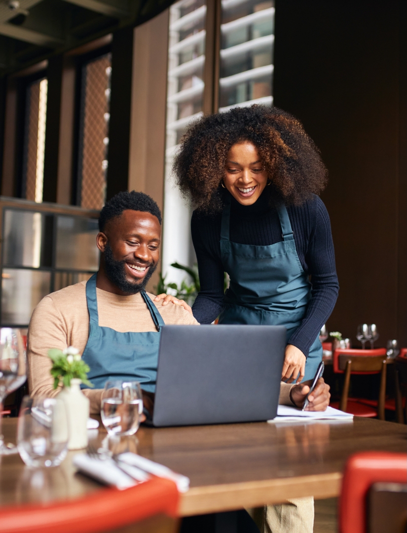 one-handsome-black-man-sit-down-and-pretty-black-woman-stand-up-next-to-it-looking-at-the-laptop-to-check-their-restaurant-marketing-plan