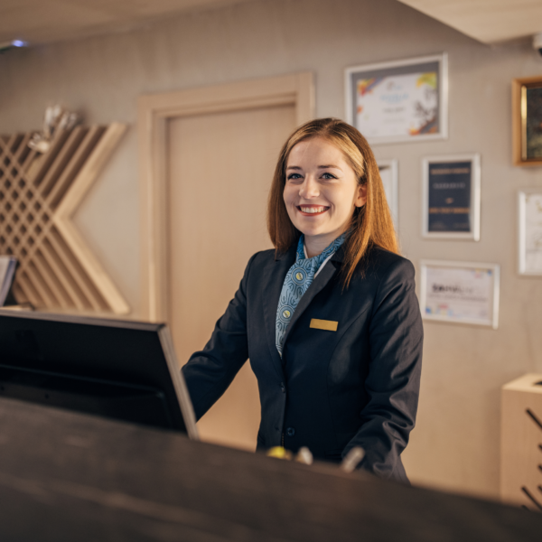 a woman wearing a hotel uniform is standing in front of the receptionist's desk serving guests who ask about artificial intelligence in hospitality industry
