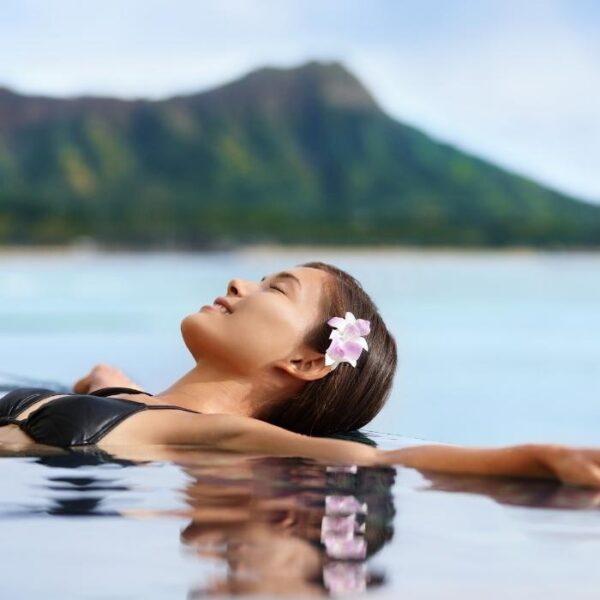 A woman with exotic brown skin and long black hair is swimming in a pool wearing a black bikini and purple flowers in her ears enjoying wellness and sustainability at Deep Roots Nusa Penida.